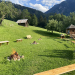 Grüne Wiese mit Lagerfeuerstelle, Holzbänken, Kindern und Blick auf Berge und Wald
