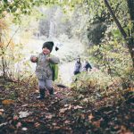 Kleines Kind mit Rucksack läuft fröhlich durch herbstlichen Wald, Familie im Hintergrund