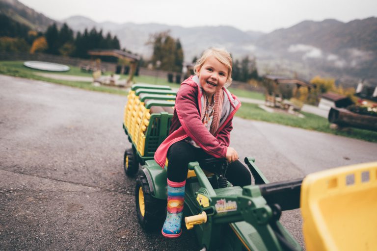 Lachendes Kind auf Spielzeugtraktor vor Alpenlandschaft am Bauernhof im Salzburger Land